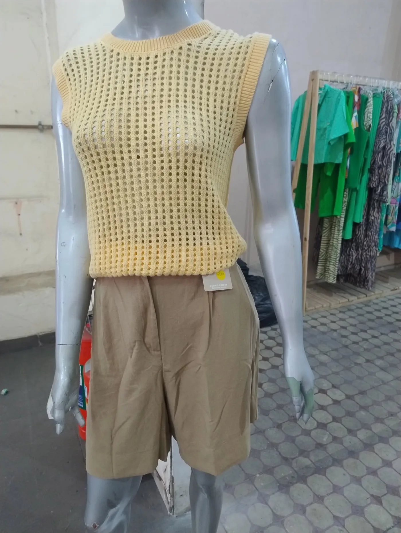 Dentro da OFF: um manequim com colete de tricô amarelo e bermuda cáqui Hering sobre o chão de ladrilho hidráulico, e araras de roupa verde e estampada ao fundo