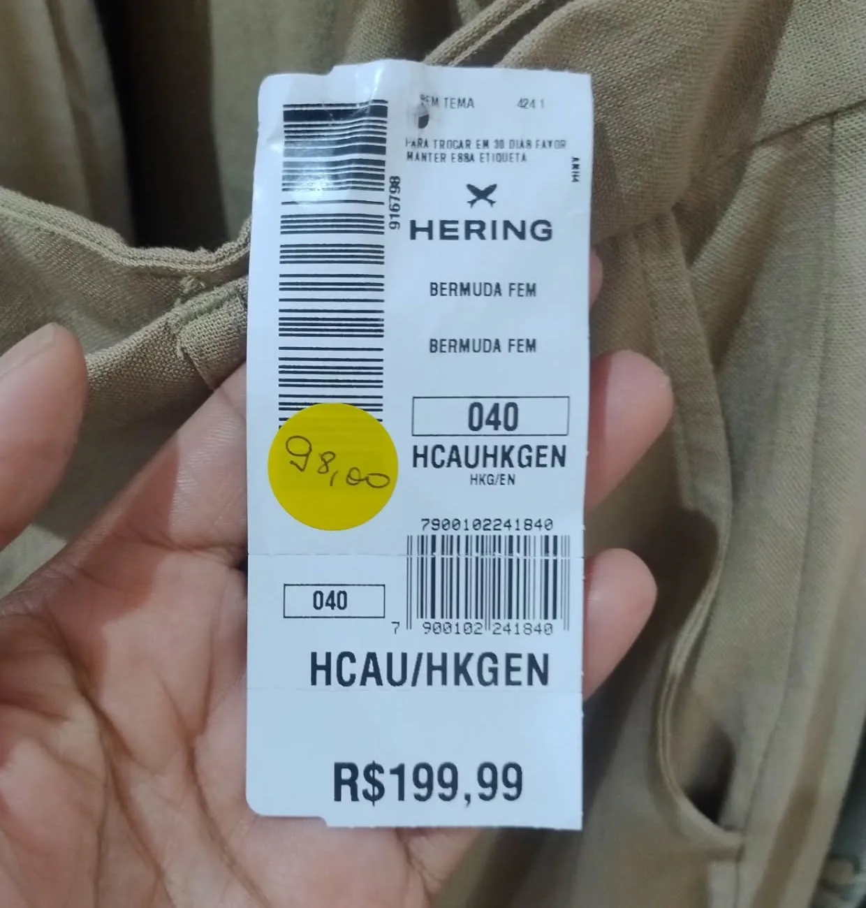 A etiqueta Hering de uma bermuda, R$ 199,99 impresso, com a bolinha amarela da OFF colada por cima e o preço novo escrito à mão